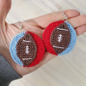 May include: A pair of crocheted earrings shaped like footballs. The earrings are red, blue, and brown with white stitching.