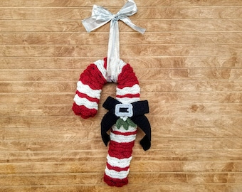 May include: A red and white striped crocheted candy cane wreath with a black bow and green holly. The wreath is hanging on a wood panel background.