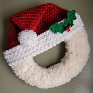 May include: A white and red crocheted Santa hat with a green holly sprig and red berries is attached to a white fluffy wreath.