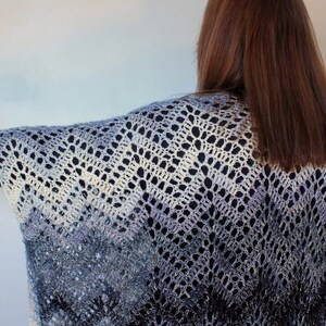 May include: A crocheted shawl with a gradient of blue, white, and gray colors. The shawl has a lacy pattern and is draped over a person's shoulders.