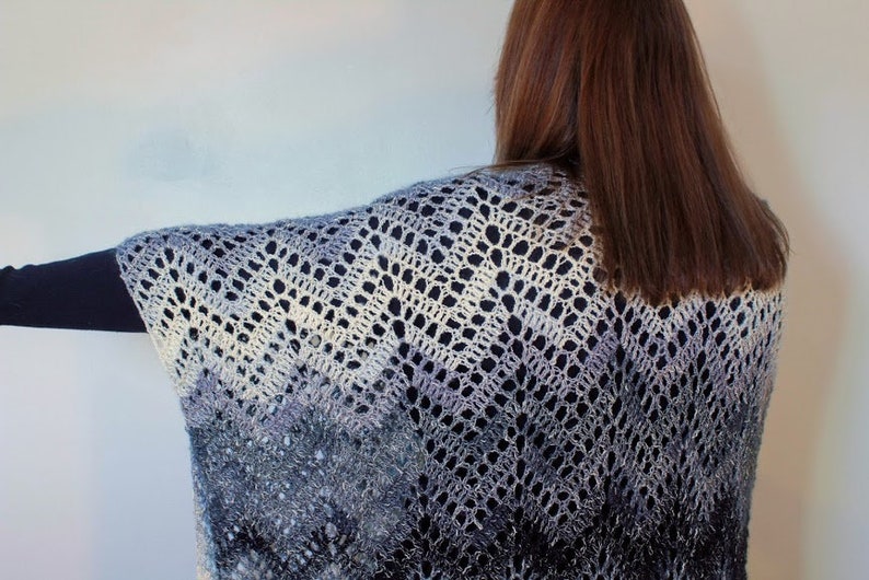 May include: A crocheted shawl with a gradient of blue, white, and gray colors. The shawl has a lacy pattern and is draped over a person's shoulders.