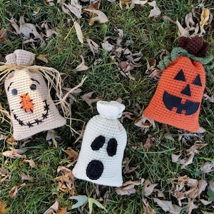 May include: Three crocheted Halloween trick-or-treat bags. One is a brown scarecrow, one is an orange pumpkin, and one is a white ghost.