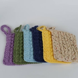 May include: Six crocheted dishcloths in various colors, including purple, green, blue, navy, yellow, and beige. The dishcloths have a textured, waffle-like pattern and are hung on a white background.