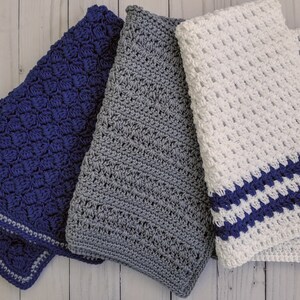 May include: Three crocheted blankets in navy blue, gray, and white with a navy blue trim. The blankets are made with a textured stitch and are folded on a white wooden surface.