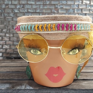 Puede incluir: Una maceta marrón con una cara pintada en ella. La cara tiene grandes gafas redondas amarillas, ojos verdes y una boca pintada de rojo. La maceta tiene una diadema trenzada marrón y blanca con cuentas de colores y una pluma amarilla.