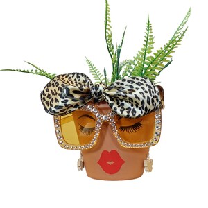 K&ouml;nnte beinhalten: Terrakotta-Blumentopf, verziert mit einem Gesicht, das eine mit Strass besetzte Sonnenbrille mit gelben Gl&auml;sern und eine Schleife im Leopardenmuster tr&auml;gt. Der Topf hat rote Lippen und falsche Wimpern. Gr&uuml;ne Pflanzen sprie&szlig;en aus der Oberseite.