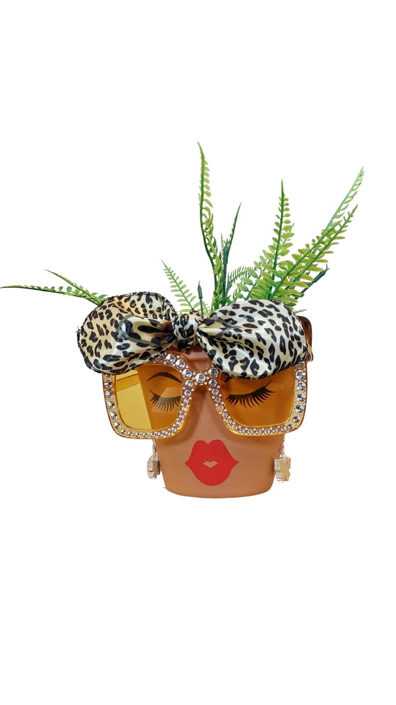 K&ouml;nnte beinhalten: Terrakotta-Blumentopf, verziert mit einem Gesicht, das eine mit Strass besetzte Sonnenbrille mit gelben Gl&auml;sern und eine Schleife im Leopardenmuster tr&auml;gt. Der Topf hat rote Lippen und falsche Wimpern. Gr&uuml;ne Pflanzen sprie&szlig;en aus der Oberseite.