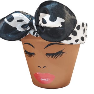 May include: A terracotta flower pot decorated with a painted face, including closed eyes, long eyelashes, and red lips. The pot is topped with a black and white polka dot bow.