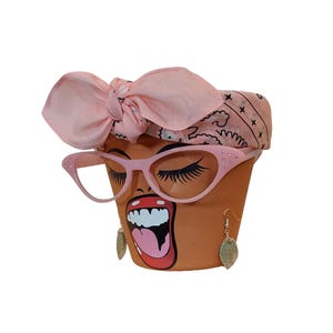 May include: A terracotta flower pot decorated with a painted face, pink cat-eye glasses, and a pink bandana. The face has long eyelashes, open mouth, and gold leaf earrings. A unique and decorative item.