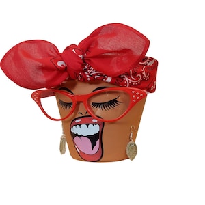 May include: A terracotta flower pot with a painted face, red glasses, and a red bandana. The face has long eyelashes, open mouth, and dangling earrings. The glasses have rhinestones.