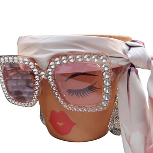May include: A decorative ceramic pot with a face design, wearing large, square, pink-tinted sunglasses encrusted with rhinestones. A pink and white scarf is tied around the pot, and a silver feather earring hangs on the side.