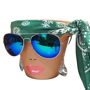 May include: A terracotta-colored flower pot decorated with a painted face, wearing blue mirrored sunglasses and a green paisley bandana. Silver feather earrings hang from the pot's rim. The pot is a decorative item.
