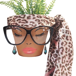 May include: A terracotta flower pot decorated with a painted face, black glasses, and turquoise earrings. The pot is adorned with a leopard print scarf and filled with green plants. A unique and stylish decorative item.