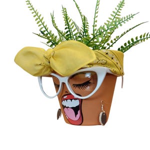 May include: A terracotta flower pot with a painted face, white cat-eye sunglasses, and a yellow bandana. The pot is decorated with a mouth graphic and earrings. Green plants sprout from the top.