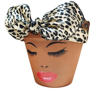 May include: A terracotta-colored flower pot with a painted face featuring closed eyes, long eyelashes, and red lips. The pot is adorned with a leopard print bow.