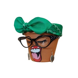 May include: A terracotta flower pot with a painted face, wearing black cat-eye glasses, a green bandana, and earrings. The face has long eyelashes and an open mouth with red lipstick. The pot is a decorative item.