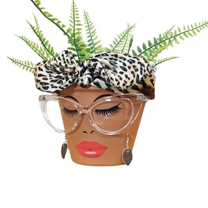 May include: A terracotta-colored flower pot decorated with a painted face, clear-framed glasses, and dangling earrings. A leopard print scarf is tied around the pot, and green fern-like plants sprout from the top.
