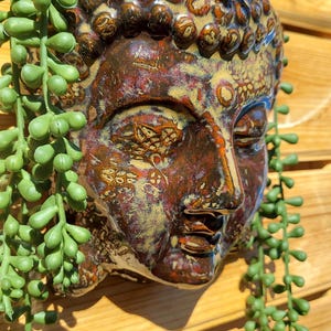May include: A ceramic Buddha head planter with a textured, multi-colored finish in shades of brown, gold, and red. The planter is filled with green, cascading string of pearls succulents. The planter has the text "Custom Pottery Sassy Soul Sister Unique Gifts".