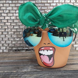 May include: A brown ceramic planter with a painted face, green bandana, and blue sunglasses. The face has a wide open mouth with white teeth and a pink tongue.