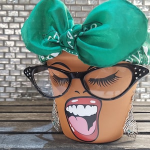 Può includere: Un vaso di ceramica marrone con un viso dipinto, un bandana verde e occhiali da sole neri a forma di occhio di gatto. Il vaso ha una grande bocca aperta con denti bianchi e una lingua rosa.