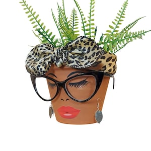 May include: A terracotta-colored flower pot decorated with a face, black eyeglasses, and silver earrings. The pot features a leopard print headscarf and is filled with green fern plants. The face has long eyelashes and red lips.