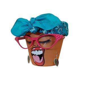 May include: A terracotta-colored flower pot with a painted face, wearing pink cat-eye glasses and teal bandana. The face has long eyelashes, a wide open mouth, and dangling leaf earrings. A unique and decorative item.