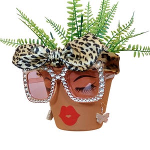 May include: A terracotta flower pot decorated with a face, wearing rhinestone-studded sunglasses with pink lenses and a leopard print bow. The pot has a red lip design and butterfly earrings, with green foliage.