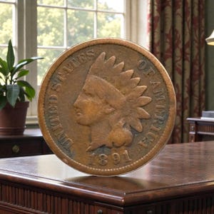 May include: A large, copper-colored Indian Head penny from 1891. The coin features a profile of a Native American in a feathered headdress and the words "United States of America." The coin is displayed on a dark wooden surface.
