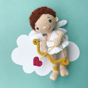 Puede incluir: Una muñeca amigurumi de crochet que representa a Cupido, con cabello castaño, sosteniendo un arco y una flecha amarillos con un corazón rojo. La muñeca lleva un vestido blanco y tiene un halo blanco.