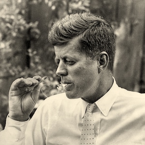 May include: A black and white photo of a man in a white shirt with a dark patterned tie, smoking a cigarette.