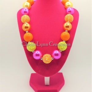 May include: A colorful chunky necklace with large beads in shades of pink, orange, and yellow. Some of the beads are solid, while others are covered in sparkly rhinestones. The necklace is displayed on a pink mannequin.