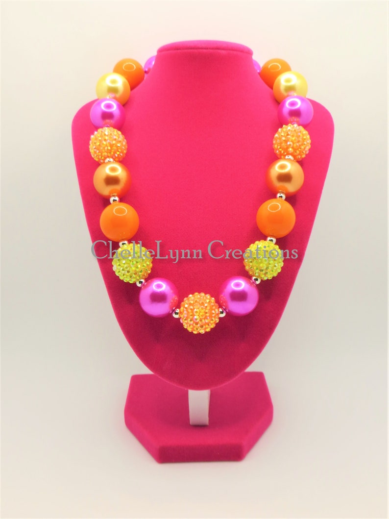 May include: A colorful chunky necklace with large beads in shades of pink, orange, and yellow. Some of the beads are solid, while others are covered in sparkly rhinestones. The necklace is displayed on a pink mannequin.