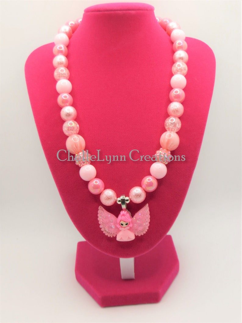 May include: Pink and white beaded necklace with a pink angel charm. The necklace is on a pink display stand.