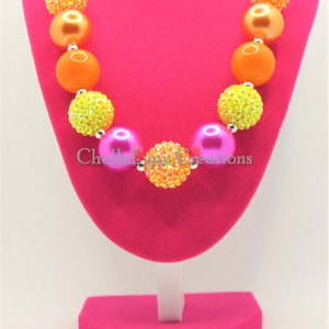 May include: A colorful chunky necklace with pink, orange, and yellow beads. The beads are decorated with rhinestones. The necklace is displayed on a pink stand.