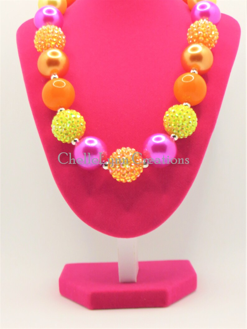 May include: A colorful chunky necklace with pink, orange, and yellow beads. The beads are decorated with rhinestones. The necklace is displayed on a pink stand.