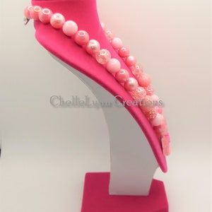 May include: A pink and white beaded necklace with a sparkly finish. The necklace is displayed on a pink velvet bust form.