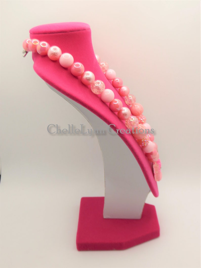 May include: A pink and white beaded necklace with a sparkly finish. The necklace is displayed on a pink velvet bust form.