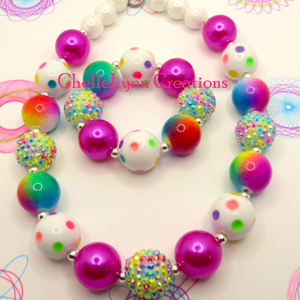 May include: A colorful beaded necklace and bracelet set. The necklace features large, round beads in white, pink, and rainbow hues, some with polka dots. The bracelet mirrors the necklace design. The text "ChelleLynn Creations" is visible.
