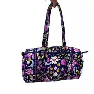 May include: A purple and green floral print duffel bag with two top handles. The bag is made of quilted fabric and has a zipper closure.