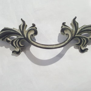 May include: Ornate drawer pull with a cream and dark gray finish. The handle features a curved design with decorative floral accents on each end. The hardware is made of metal and is designed to be mounted on a drawer or cabinet.