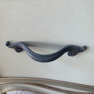 May include: Close-up of a drawer with a dark, curved handle. The drawer is painted a pale green with a textured surface. The handle has decorative scroll details on each end and is mounted on a cream-colored frame with gold trim.