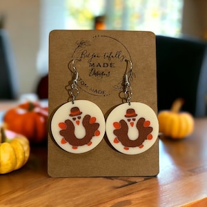 May include: A pair of white, round earrings with a brown turkey design and orange accents. The earrings have silver hooks and are displayed on a brown card with the text "Bel-air-tifully MADE Designs CUSTOM MADE".