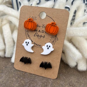 May include: A set of six Halloween-themed stud earrings. Two orange pumpkin earrings, two white ghost earrings, and two black bat earrings. The earrings are displayed on a brown card with the text "Custom Made Designs".