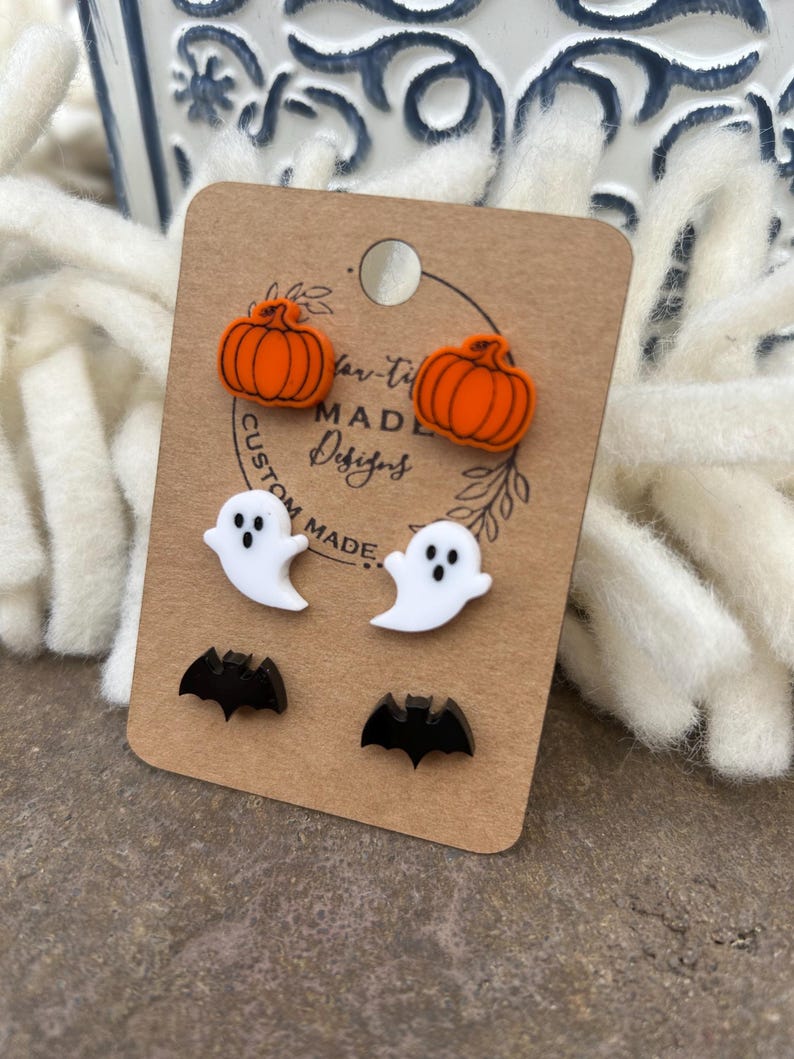 May include: A set of six Halloween-themed stud earrings. Two orange pumpkin earrings, two white ghost earrings, and two black bat earrings. The earrings are displayed on a brown card with the text "Custom Made Designs".