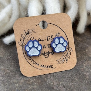 May include: A pair of blue and silver paw print stud earrings. The earrings are on a brown card with the text "Bear-Tiffle Designs Custom Made..."
