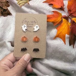 May include: A set of Halloween-themed earrings featuring a pair of white ghosts, a pair of orange pumpkins, and a pair of black bats. The earrings are displayed on a brown card with the text "Be You Tifully Made Designs" printed on it.