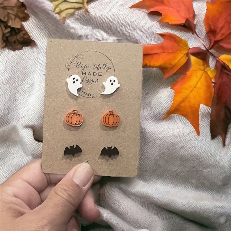 May include: A set of Halloween-themed earrings featuring a pair of white ghosts, a pair of orange pumpkins, and a pair of black bats. The earrings are displayed on a brown card with the text "Be You Tifully Made Designs" printed on it.