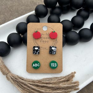 May include: A set of four pairs of earrings featuring a red apple, a black and white checkered pattern, and green circles with the letters "ABC" and the numbers "123" in white. The earrings are displayed on a brown card with the text "Far-Tiff Made Designs".