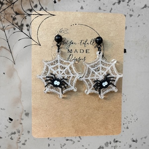 May include: A pair of black spider web earrings with glitter and a black spider on each web. The earrings are on a brown card with the text "Belfort-tifally MADE Designs".
