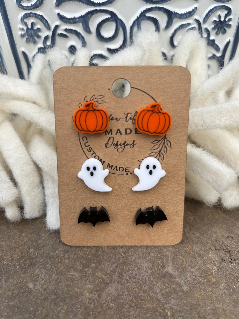 May include: A set of six Halloween-themed earrings. The earrings are made of acrylic and feature pumpkins, ghosts, and bats. The earrings are displayed on a brown card with the text "Tan-Tif Made Designs" and "Custom Made".